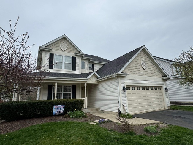 A front-facing, ground-level view of a two-story beige house with a newly installed dark grey asphalt shingle roof. The image captures the front of the home, including a two-car garage, front porch, and black shutters, with a lawn sign visible in the foreground.